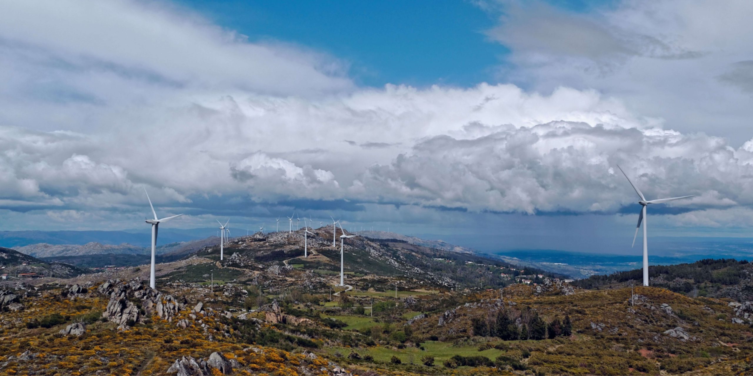 Parque eólico numa zona montanhosa de Portugal, com várias turbinas a produzir energia renovável sob um céu nublado, representando o consumo de eletricidade e a transição energética. 