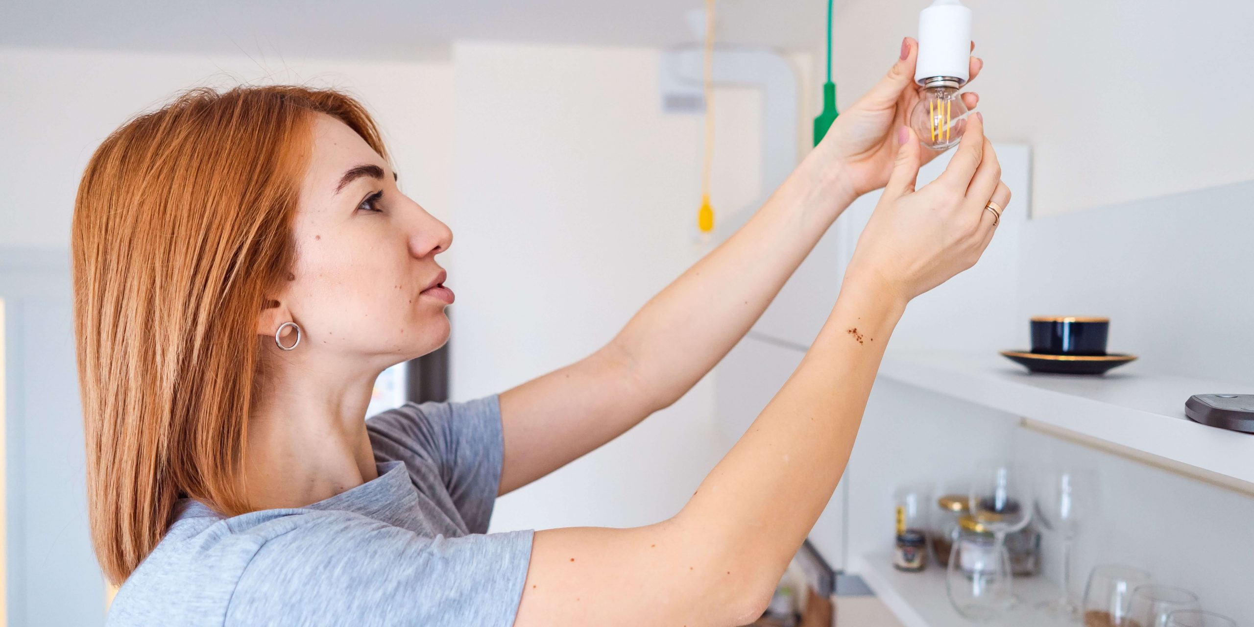 Mulher a trocar uma lâmpada em casa, num artigo sobre potência contratada, consumo de eletricidade e formas de poupar na fatura da luz.