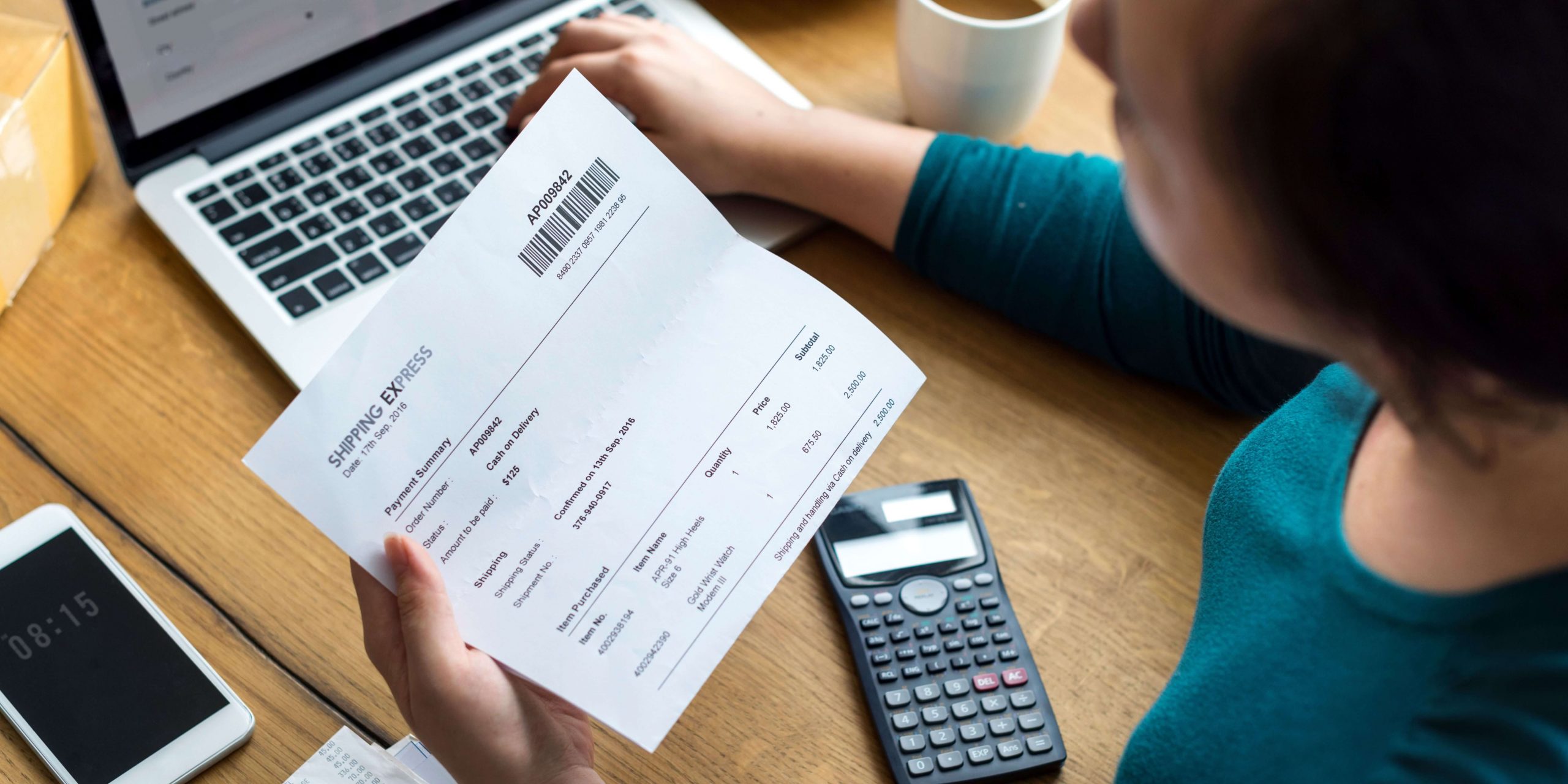 Pessoa a analisar uma fatura em papel junto a um computador portátil, calculadora e telemóvel, representando a gestão de despesas domésticas, controlo de pagamentos e comparação de custos para poupar na eletricidade.