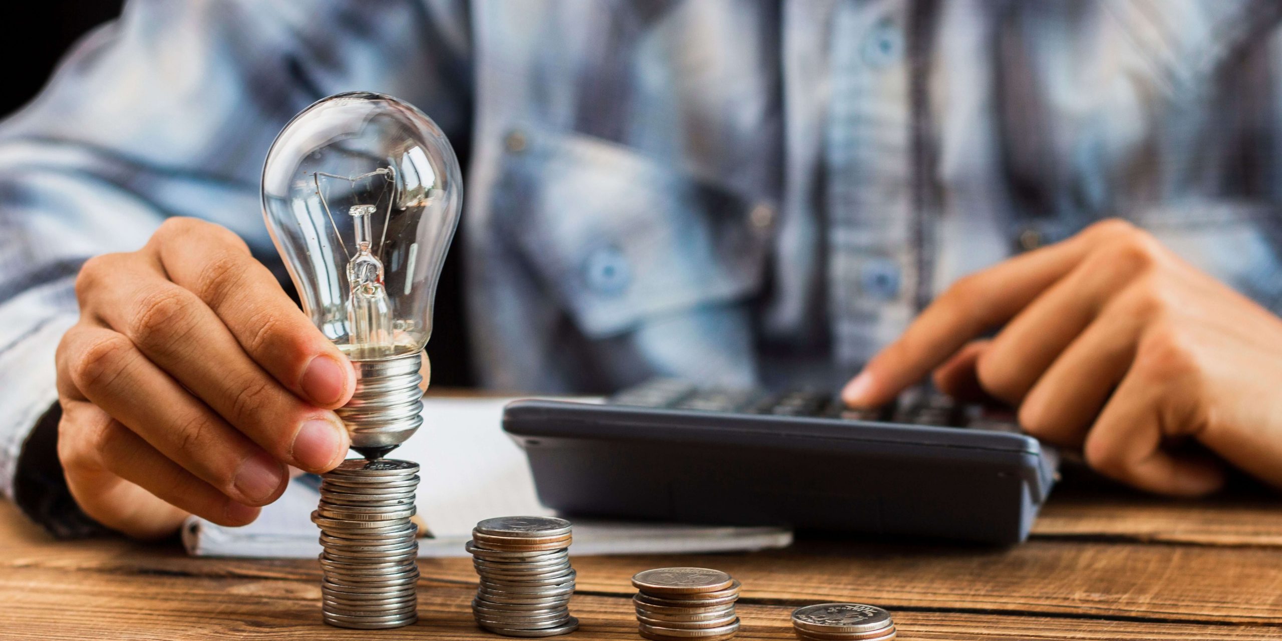Pessoa segura uma lâmpada sobre moedas enquanto utiliza uma calculadora, representando a comparação entre preço fixo e indexado na eletricidade e a poupança nas faturas.