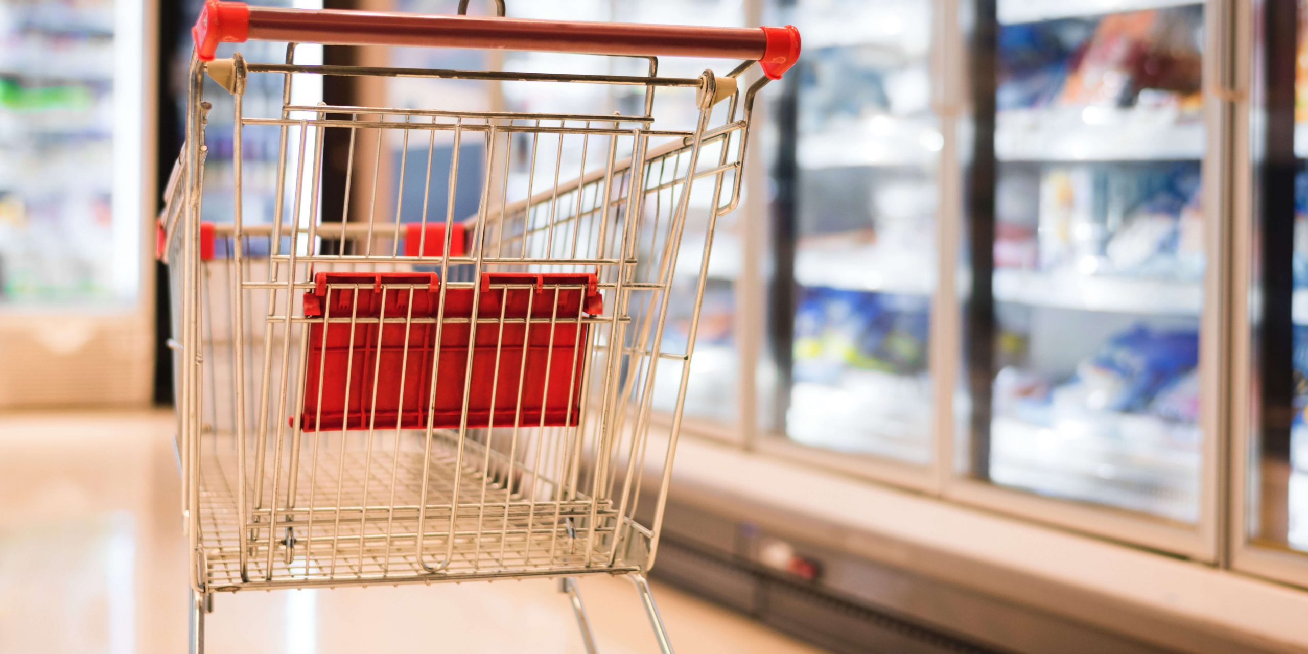 Carrinho de supermercado vazio junto a prateleiras refrigeradas, representando compras alimentares, apoio alimentar e Cartão Social eletrónico em Portugal.